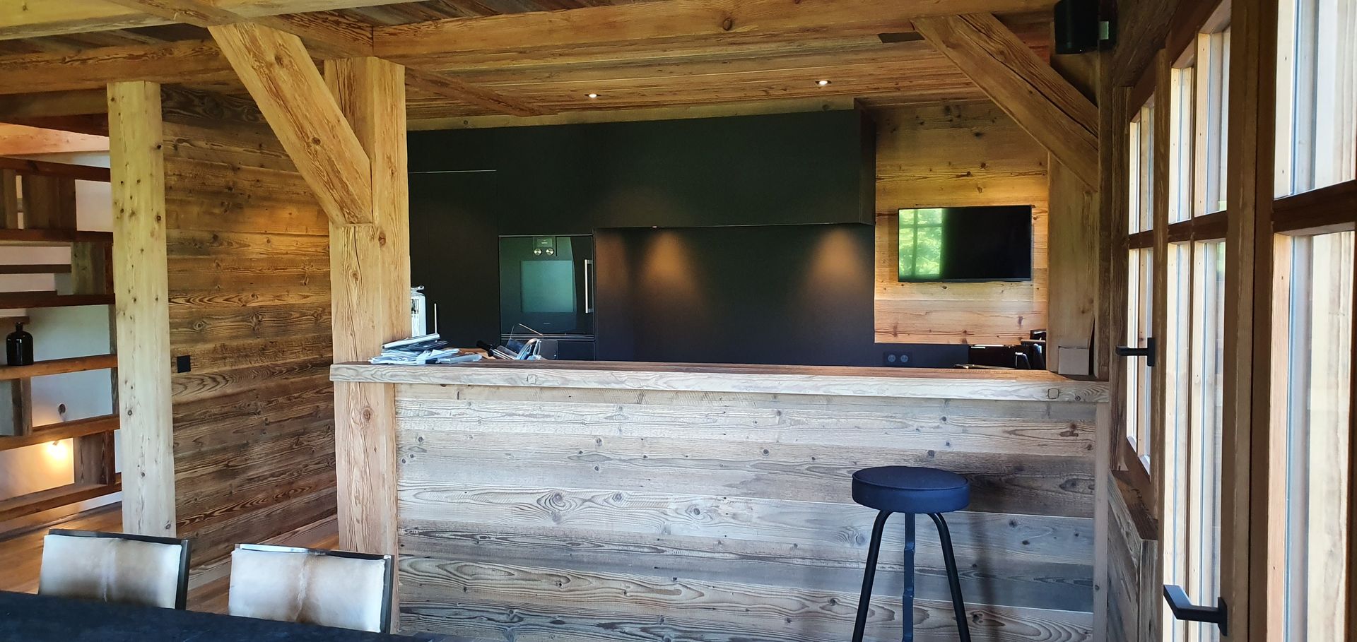 Bar rustique avec poutres et lambris en bois, mur d'accent sombre et télévision ; un tabouret est installé au bar.