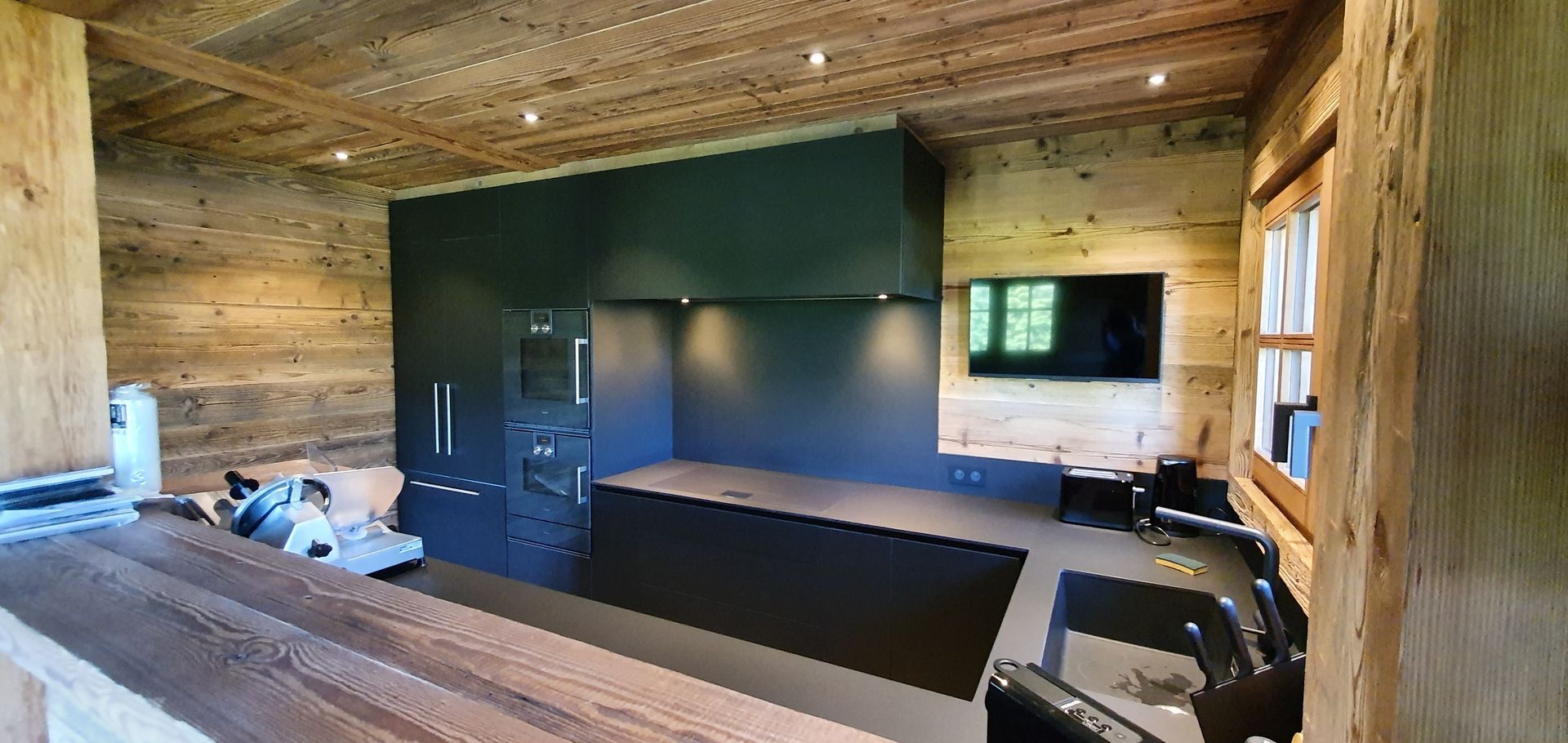 Cuisine moderne avec placards noirs et murs en bois. Il y a une télévision au mur.