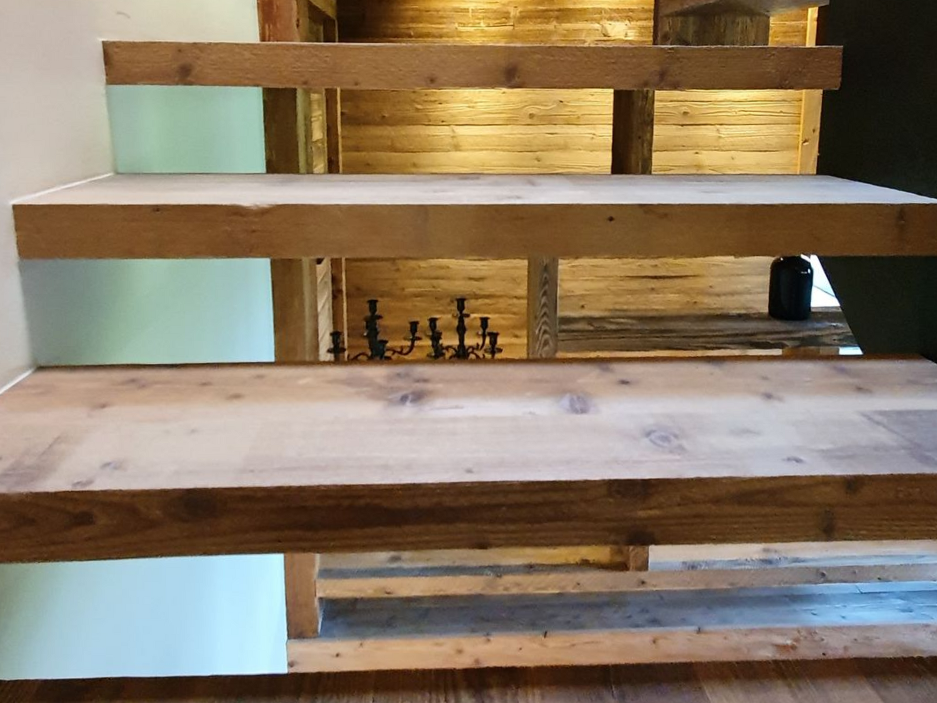 Escalier en bois aux veines apparentes, menant à l'étage ; bibliothèque visible en arrière-plan.