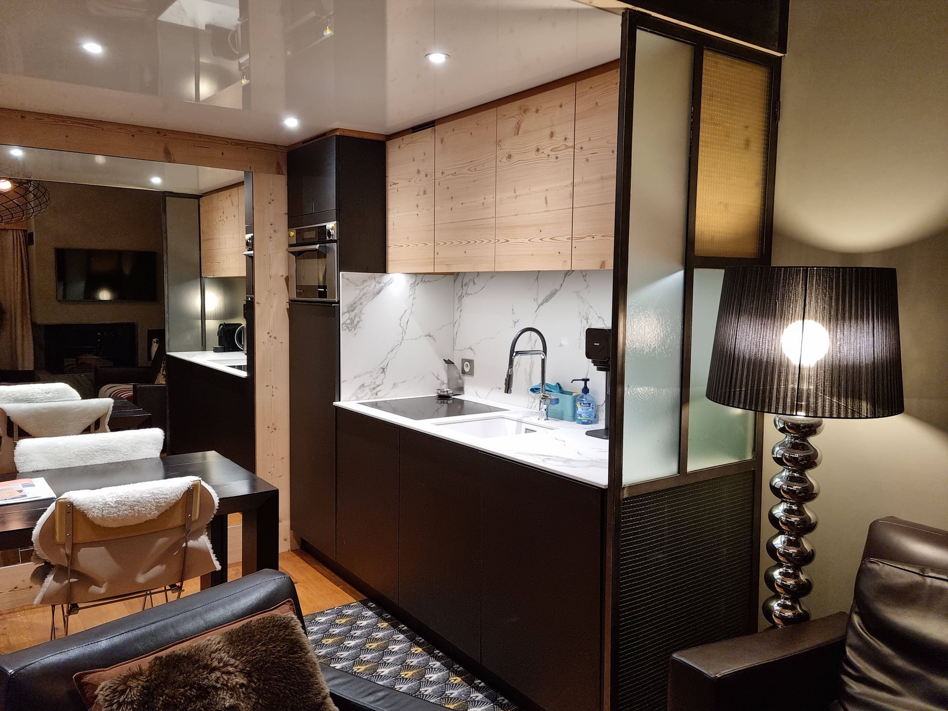 Cuisine chaleureuse avec lambris, placards foncés et plans de travail en marbre, séparée du séjour par une paroi de verre.