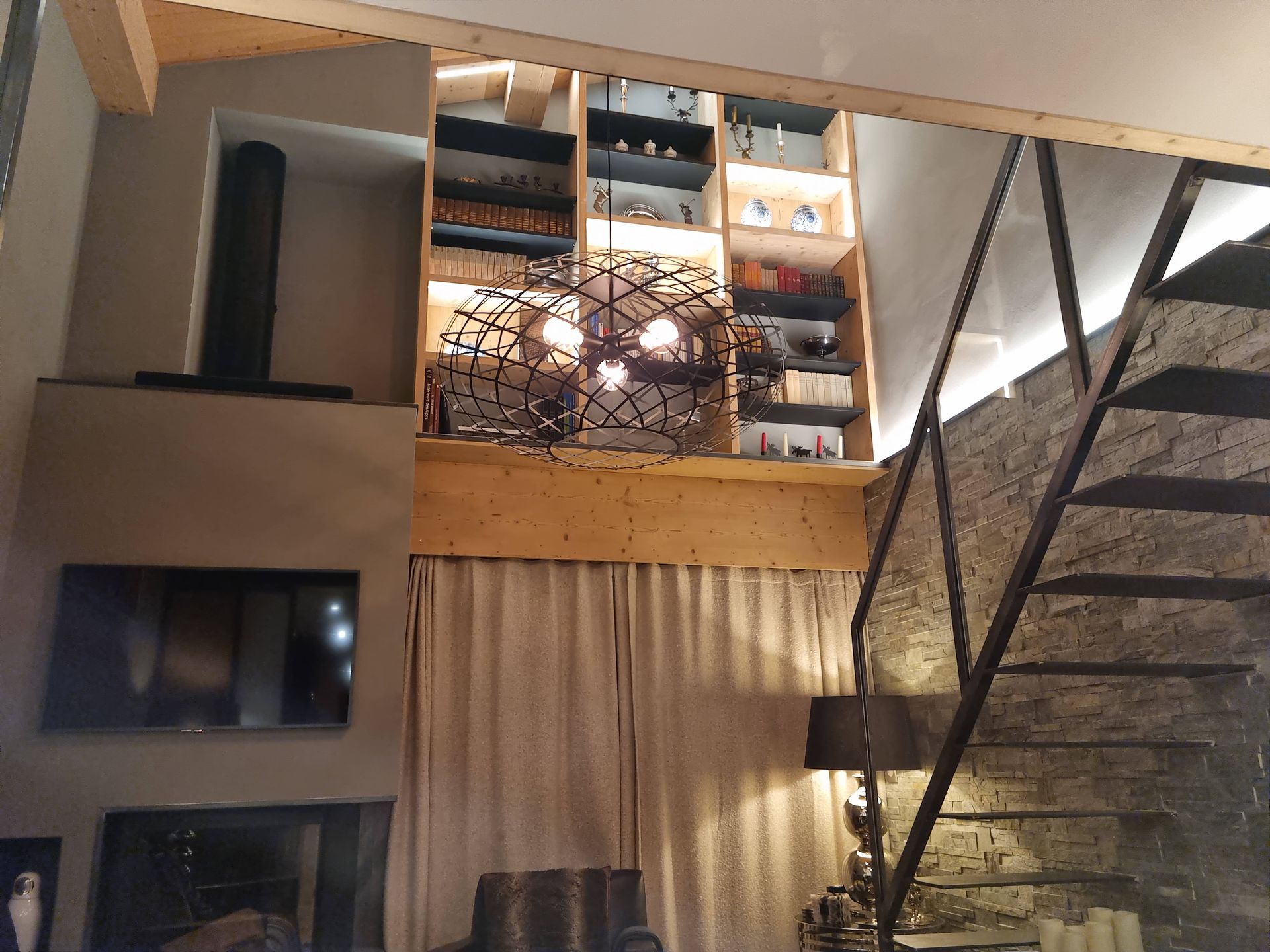 Intérieur avec escalier, étagères, cheminée et lustre. Éléments en bois, mur en pierre et fenêtre à rideaux.
