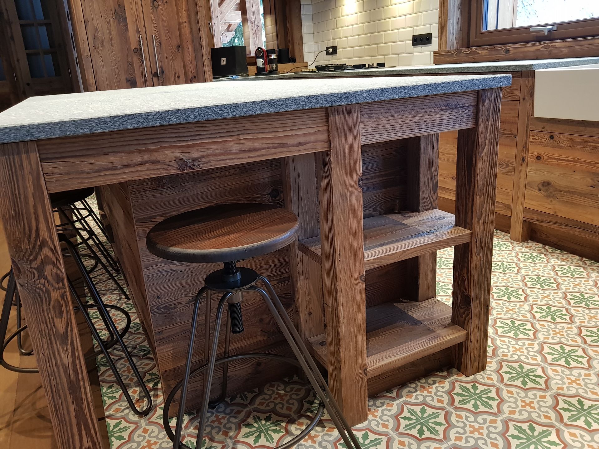 Îlot de cuisine en bois avec plan de travail gris, tabouret et étagères sur un sol carrelé à motifs.