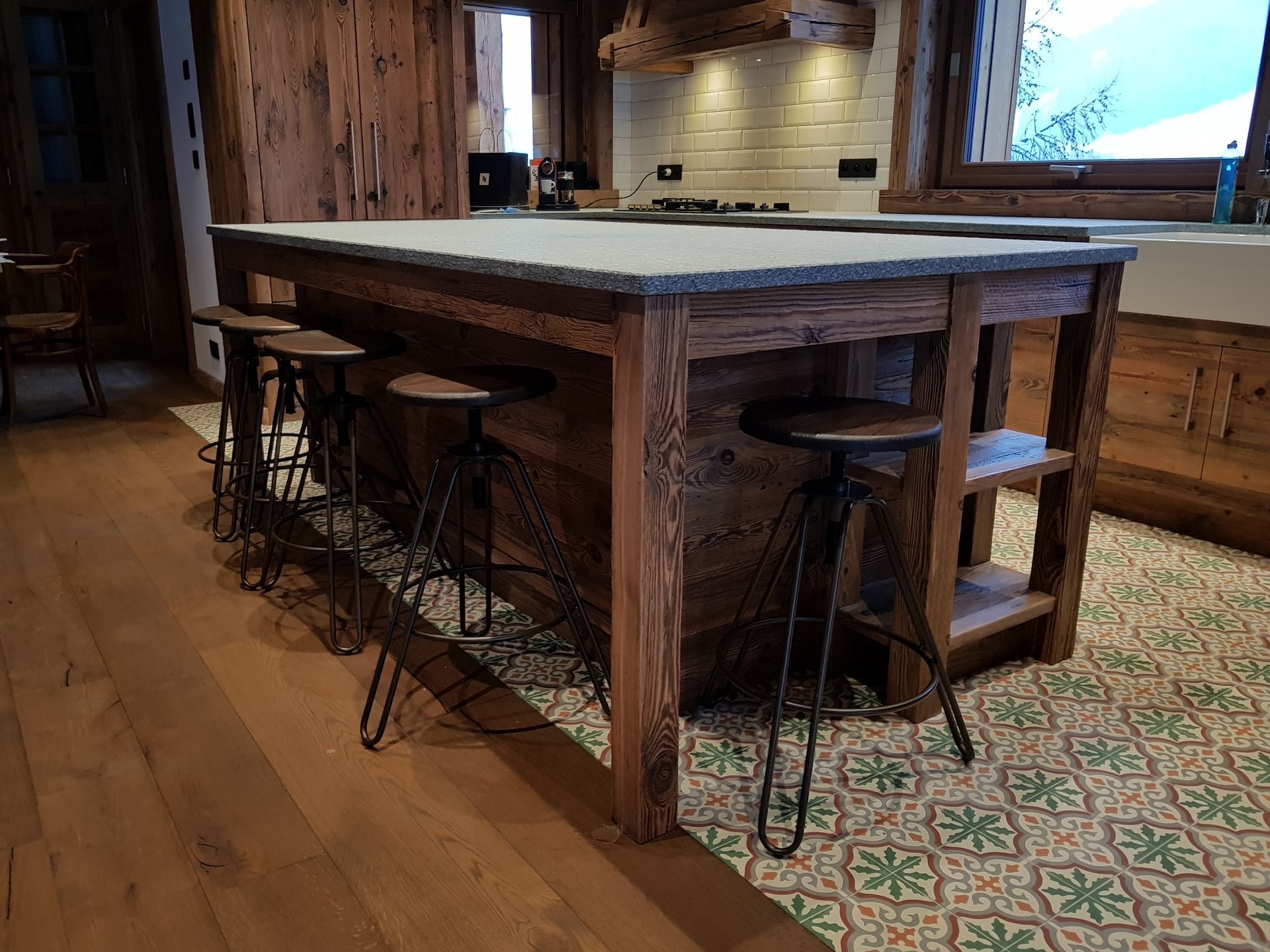 Îlot de cuisine avec comptoir en pierre, structure en bois et tabourets. Le sol est recouvert de carrelage à motifs et de parquet.