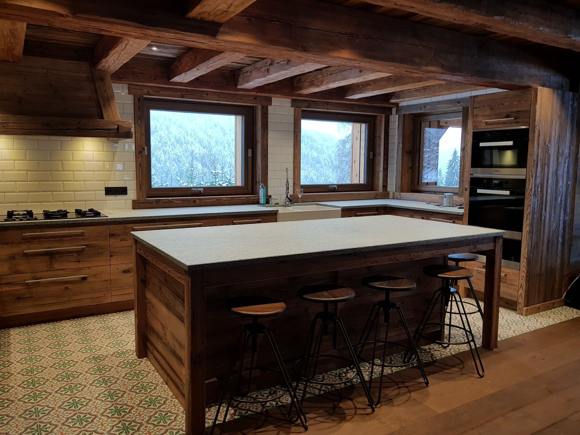 Cuisine rustique avec placards en bois, îlot central avec tabourets et fenêtres donnant sur un paysage enneigé.