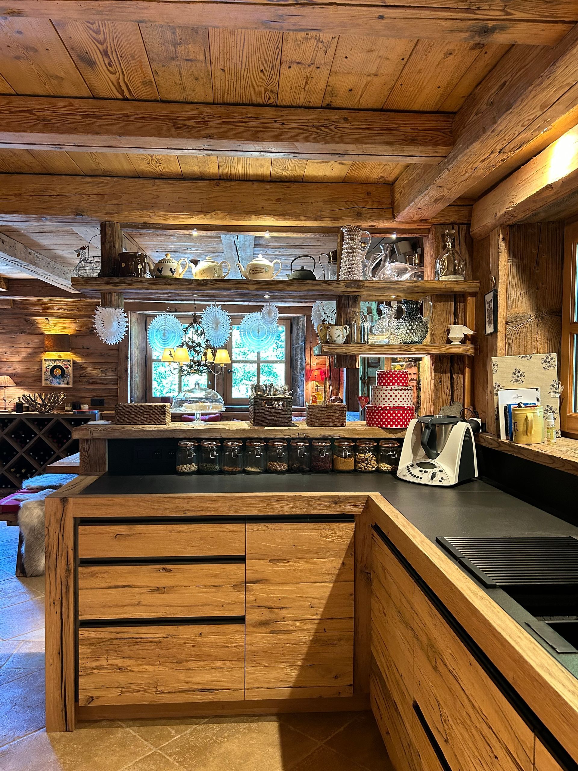 Cuisine en bois avec étagères présentant la vaisselle, plan de travail avec bocaux et appareils électroménagers, et une fenêtre.