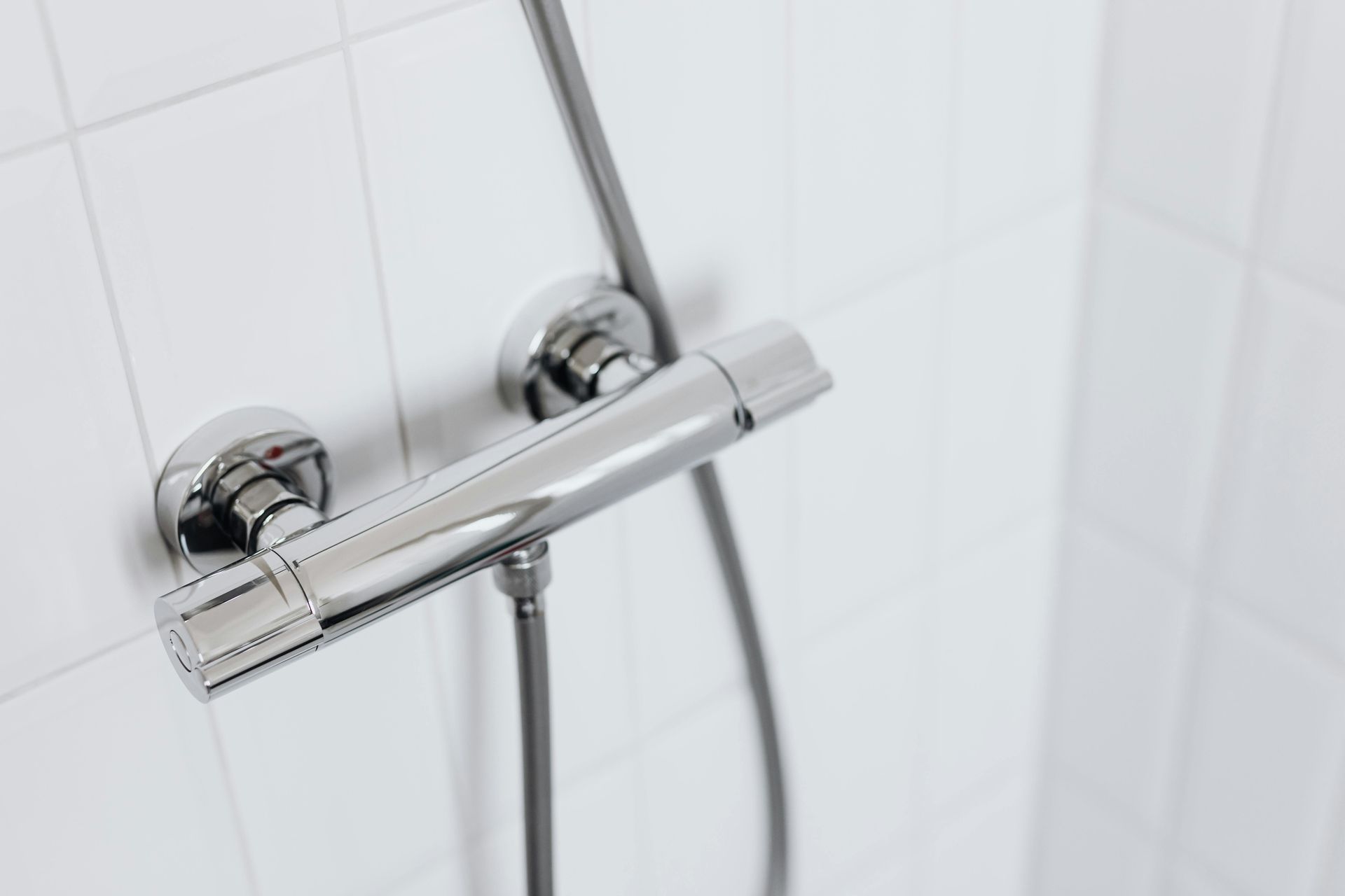 Shiny chrome shower fixtures on a white tiled wall, including a temperature control valve and flexible hose.