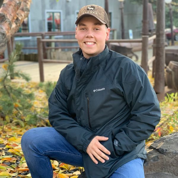Young man in a Columbia jacket and baseball cap kneels outdoors, smiling. He's surrounded by autumn leaves and a rustic setting.