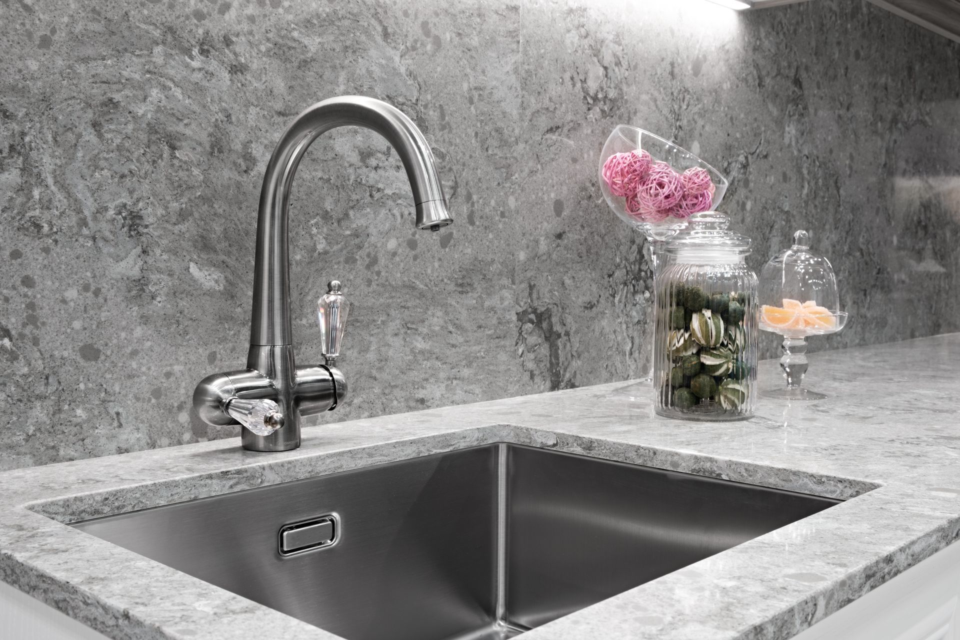 Stainless steel kitchen sink with faucet and countertop, against a gray stone backsplash, with decorative jars.