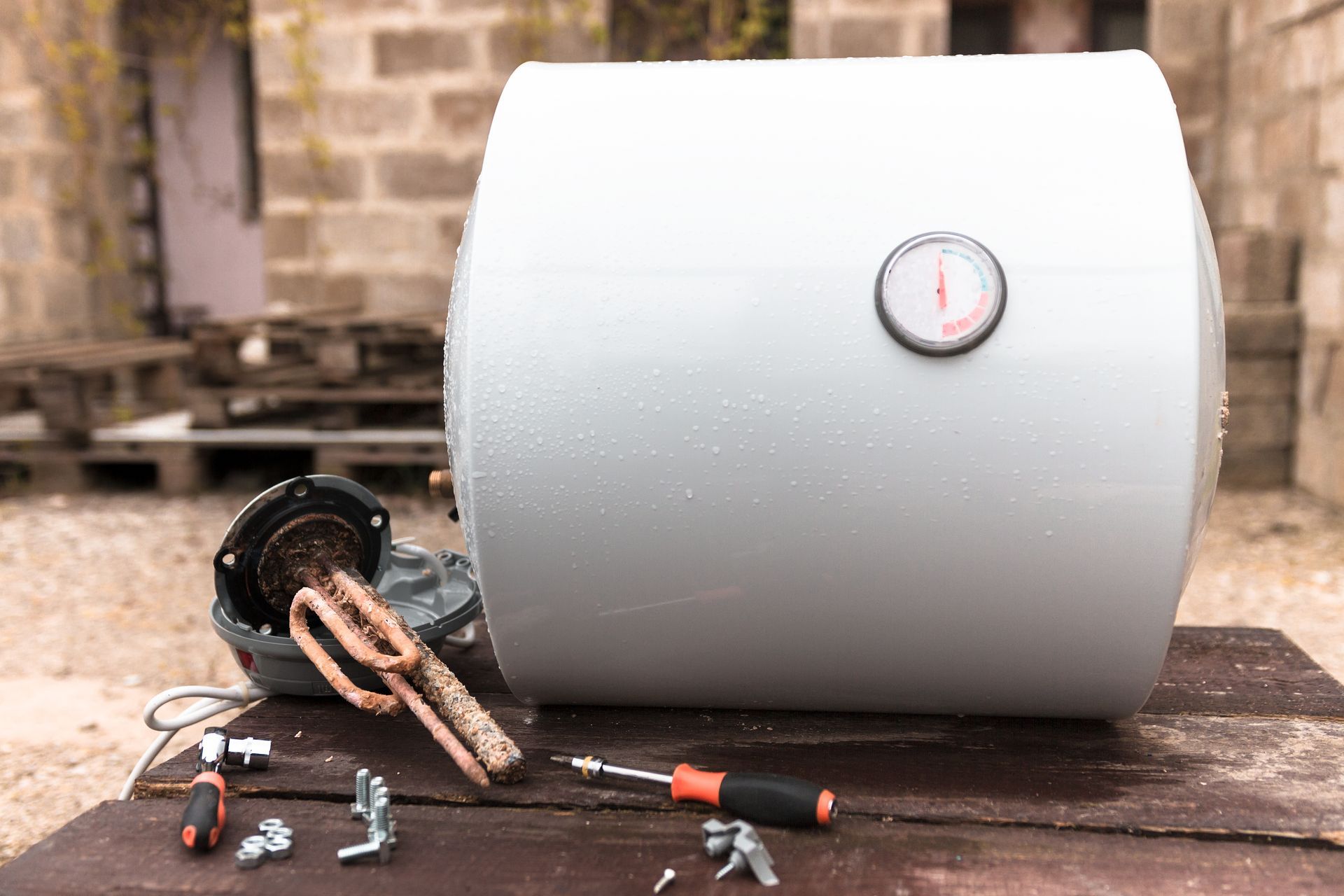 Disassembled water heater on a wooden table outdoors, with its components and a screwdriver.