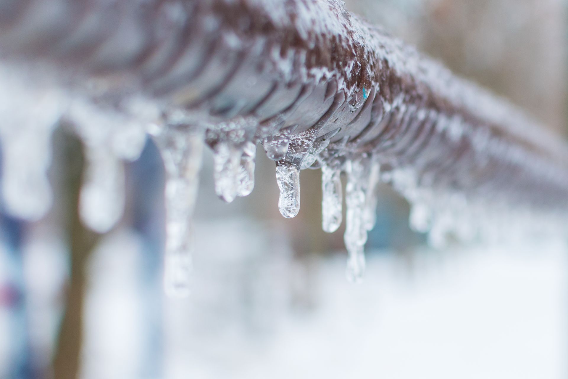 Icicles hanging from a brown metal pipe, blurry winter backdrop.