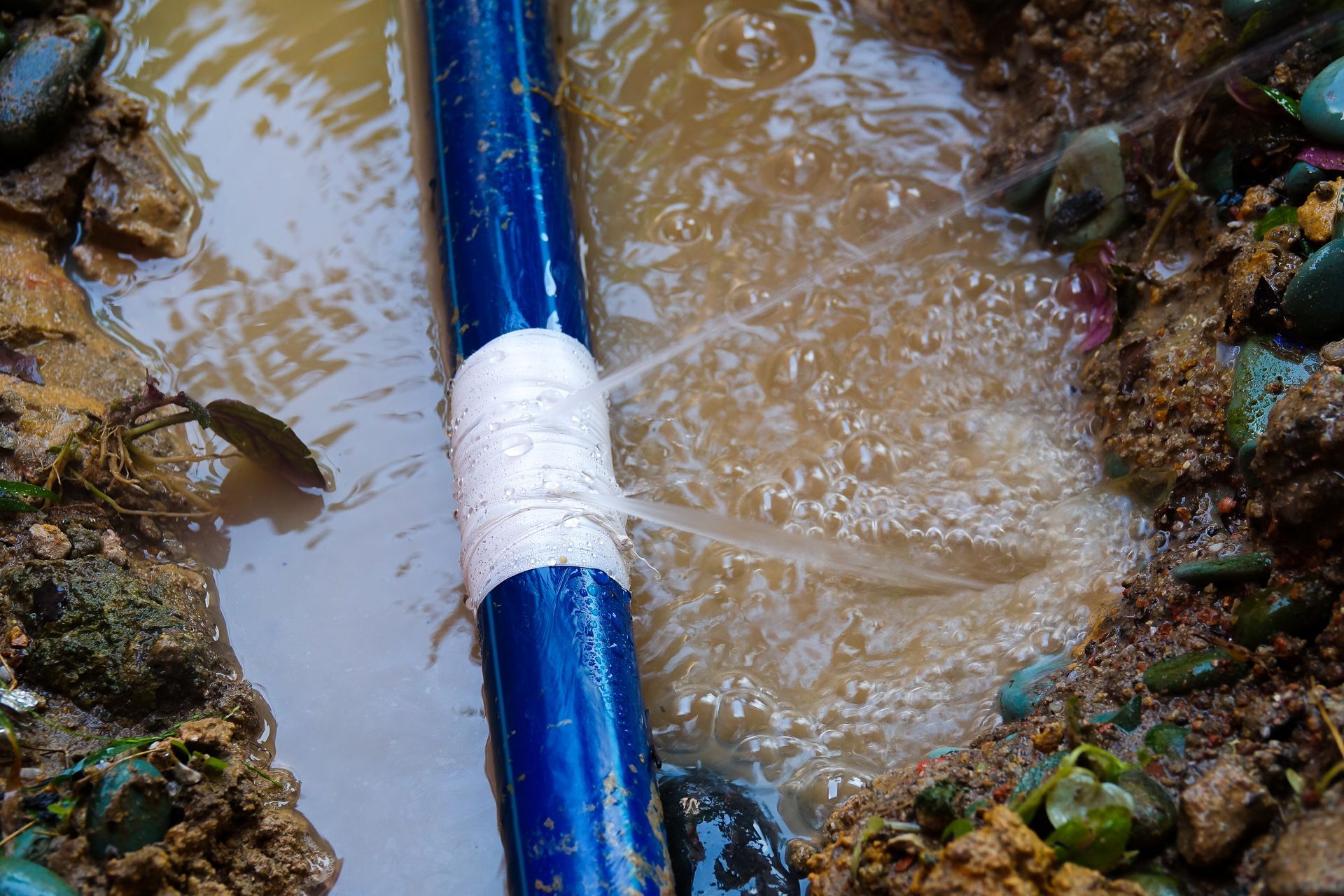 Leaking blue pipe wrapped with white tape; water spurting into muddy ground.