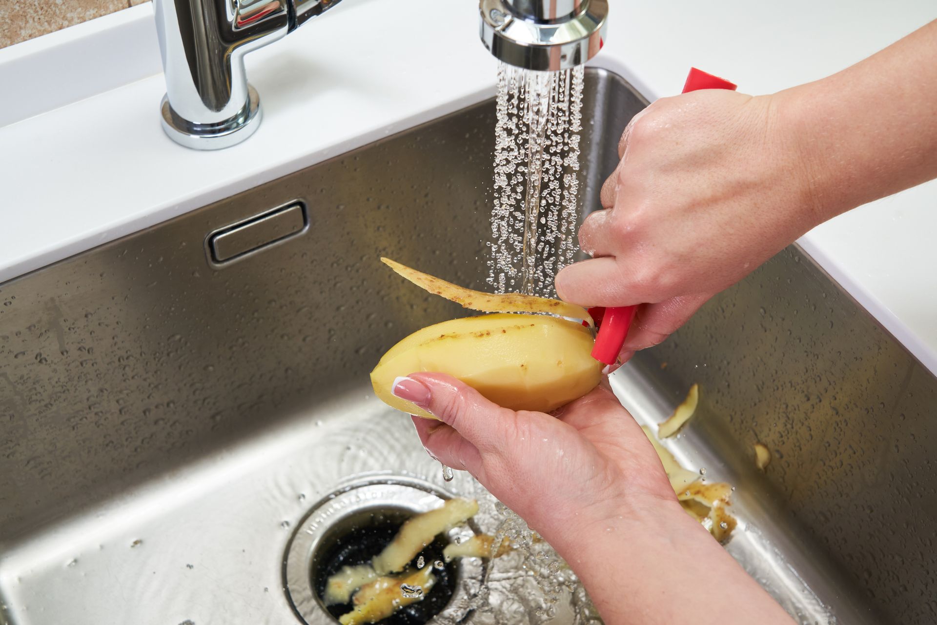 Person peeling a potato in a kitchen sink with running water, using a red peeler.