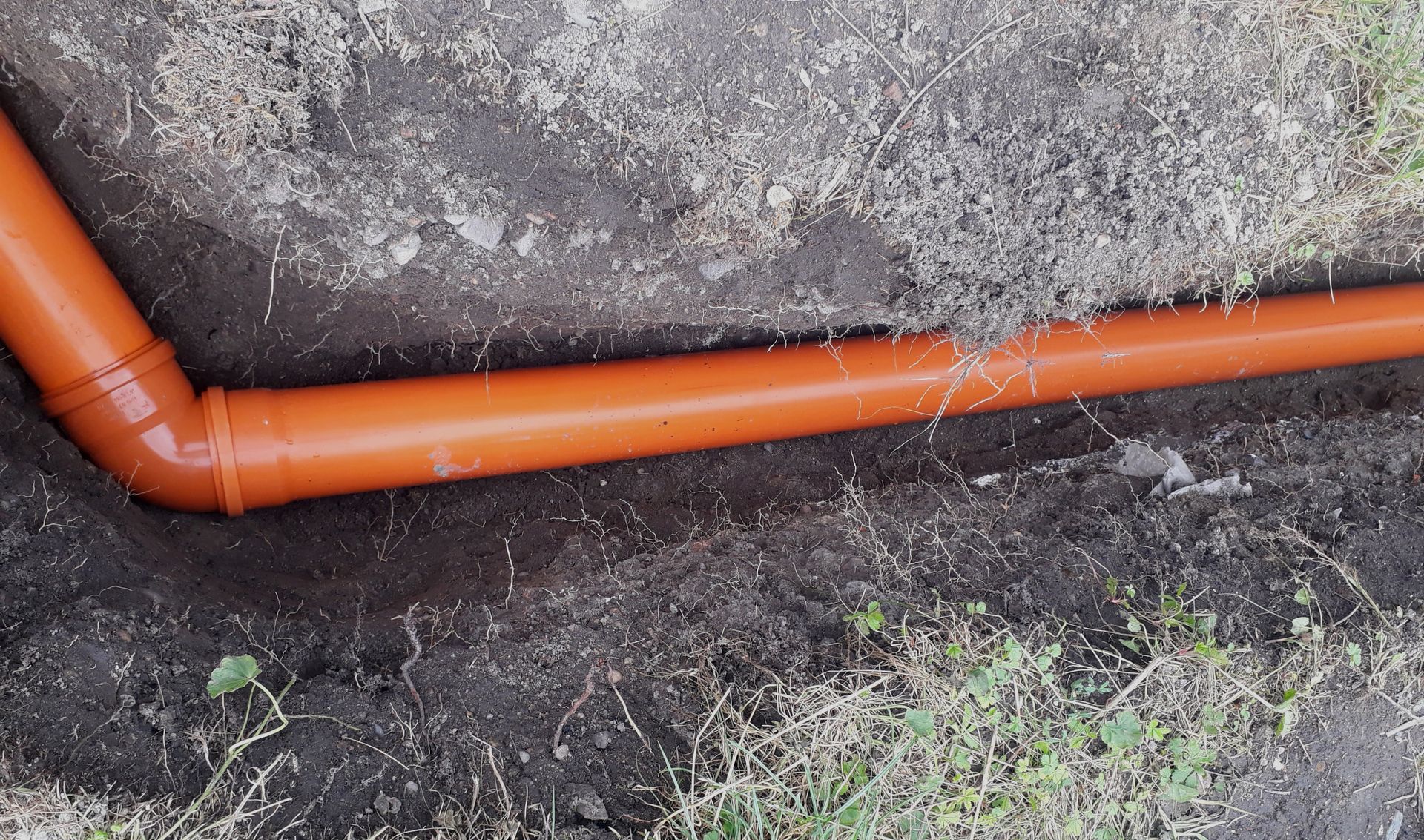 Orange plastic drainage pipe laid in a shallow trench in the ground.