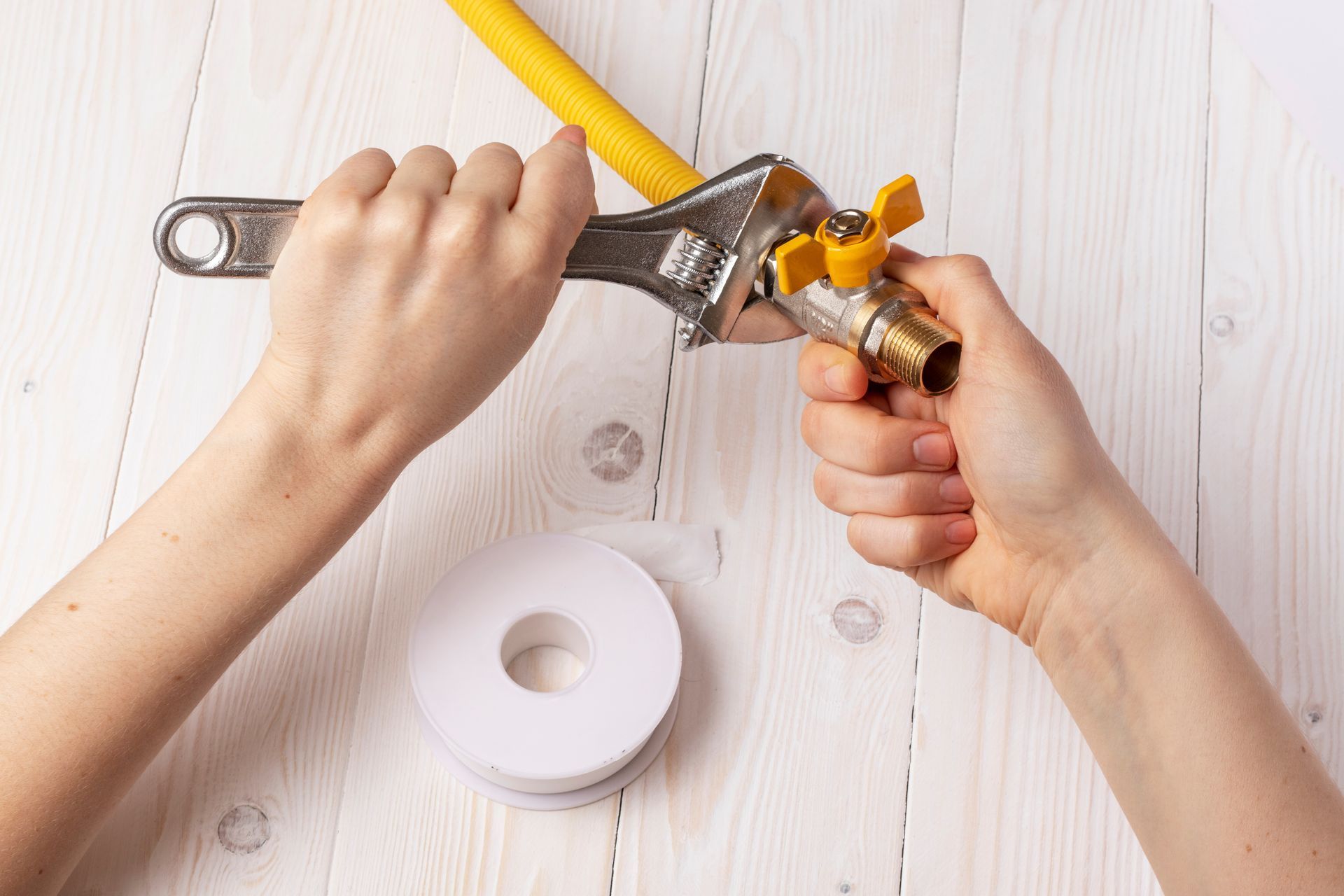 Hands using a wrench to tighten a valve on a yellow gas line, with Teflon tape on a white wooden surface.