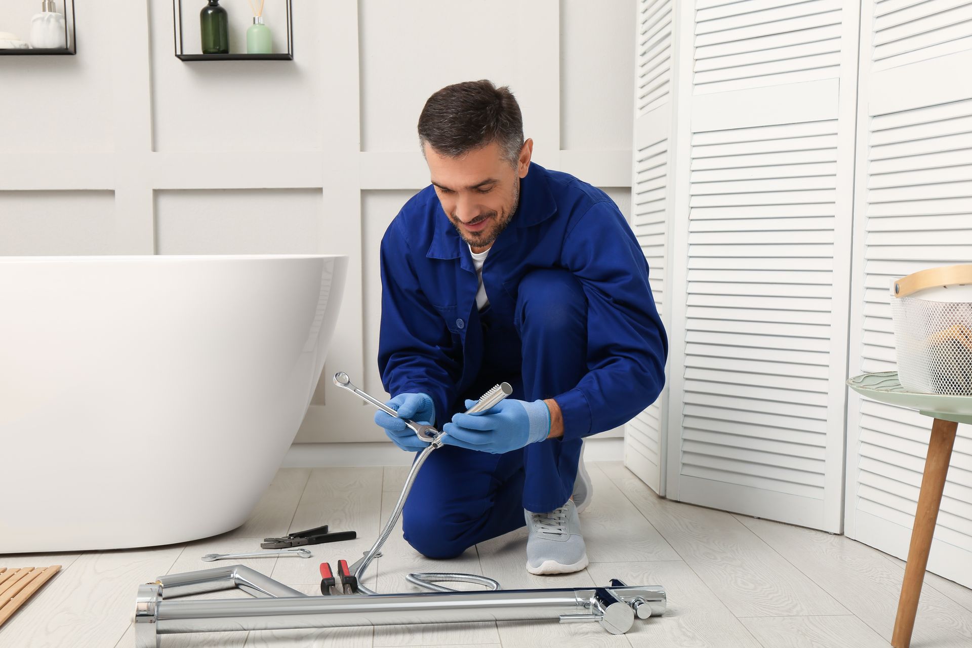 Plumber in blue coveralls and gloves repairing plumbing in a bathroom with a white tub and shoji screen.