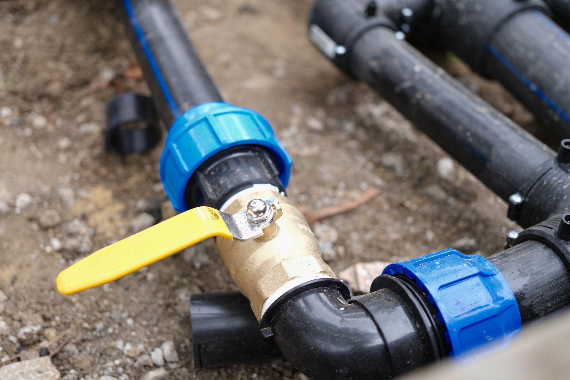 Yellow-handled valve in brass, blue-capped black pipe fittings. Outdoor setting.