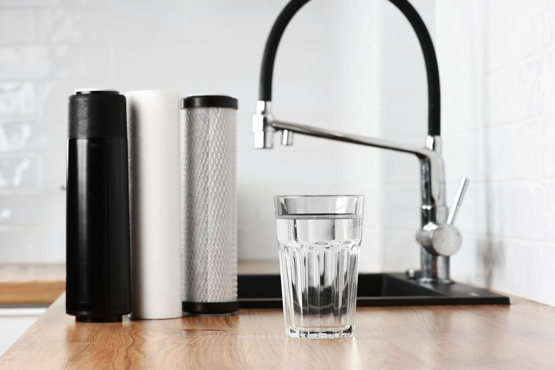Water filter cartridges next to a faucet and glass of water on a wooden counter.