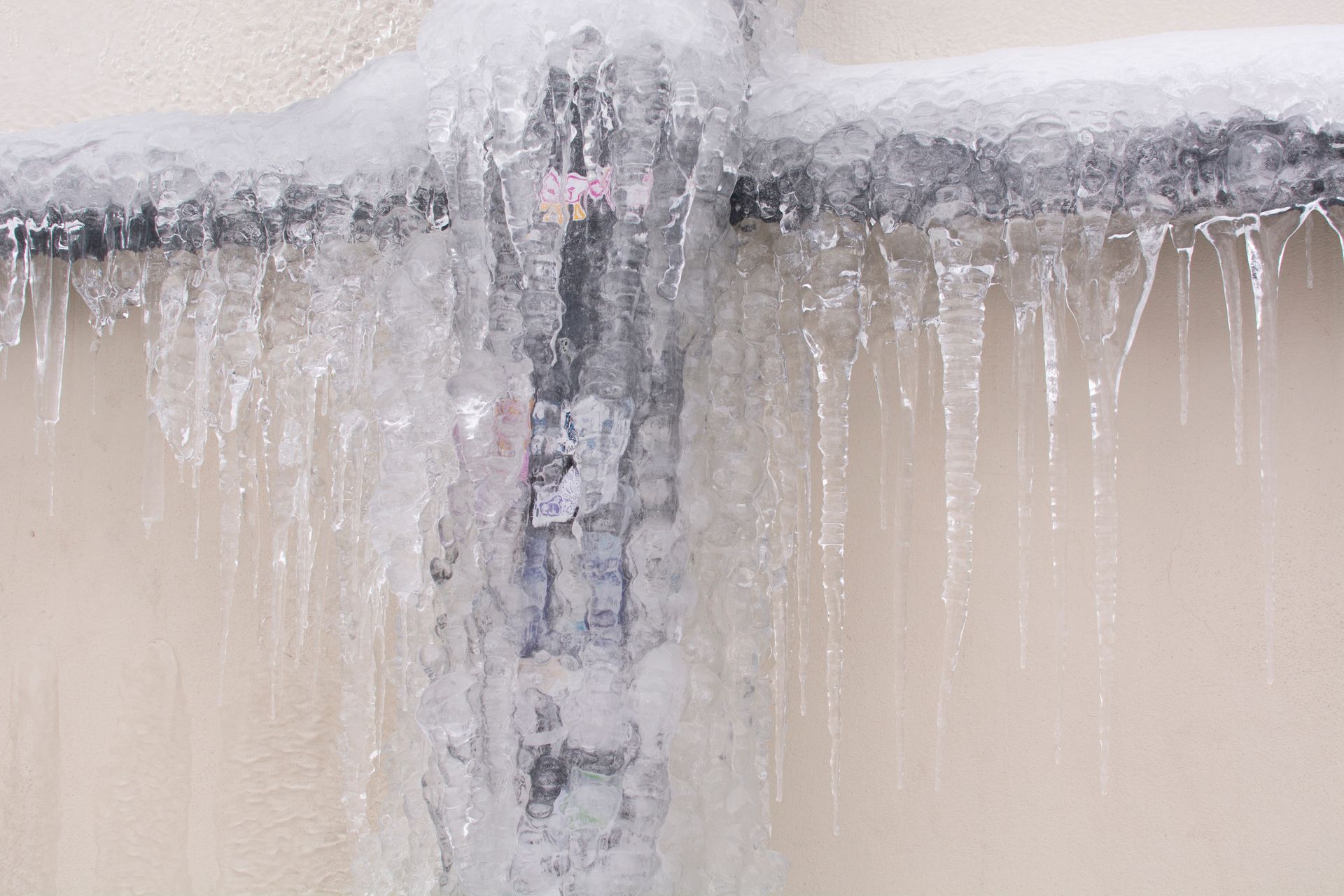 Ice encases outdoor pipes. Long icicles hang from the frozen metal against a light-colored wall.