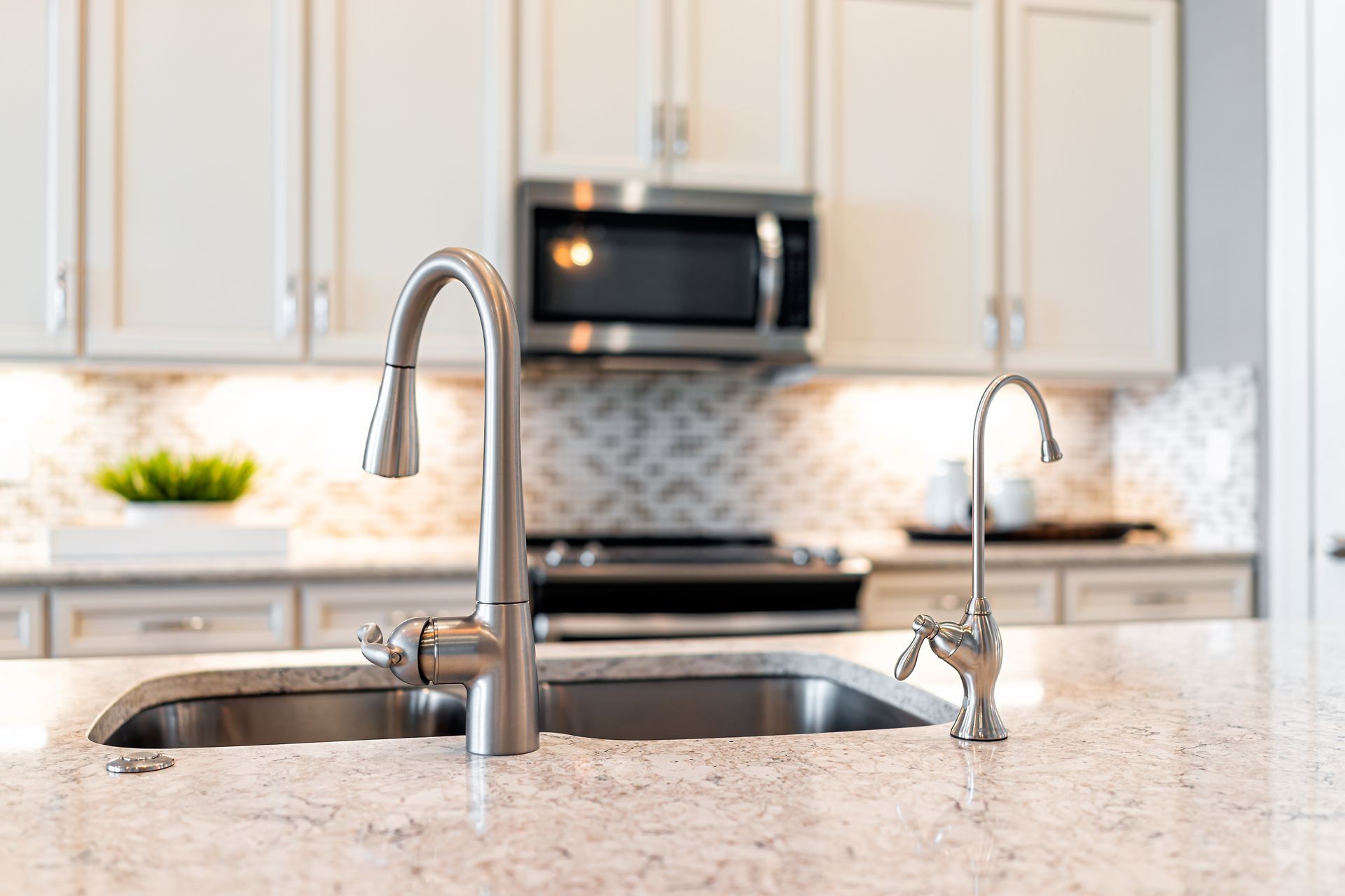 Stainless steel kitchen sink with two faucets, set in a white countertop. Cabinets and microwave in background.