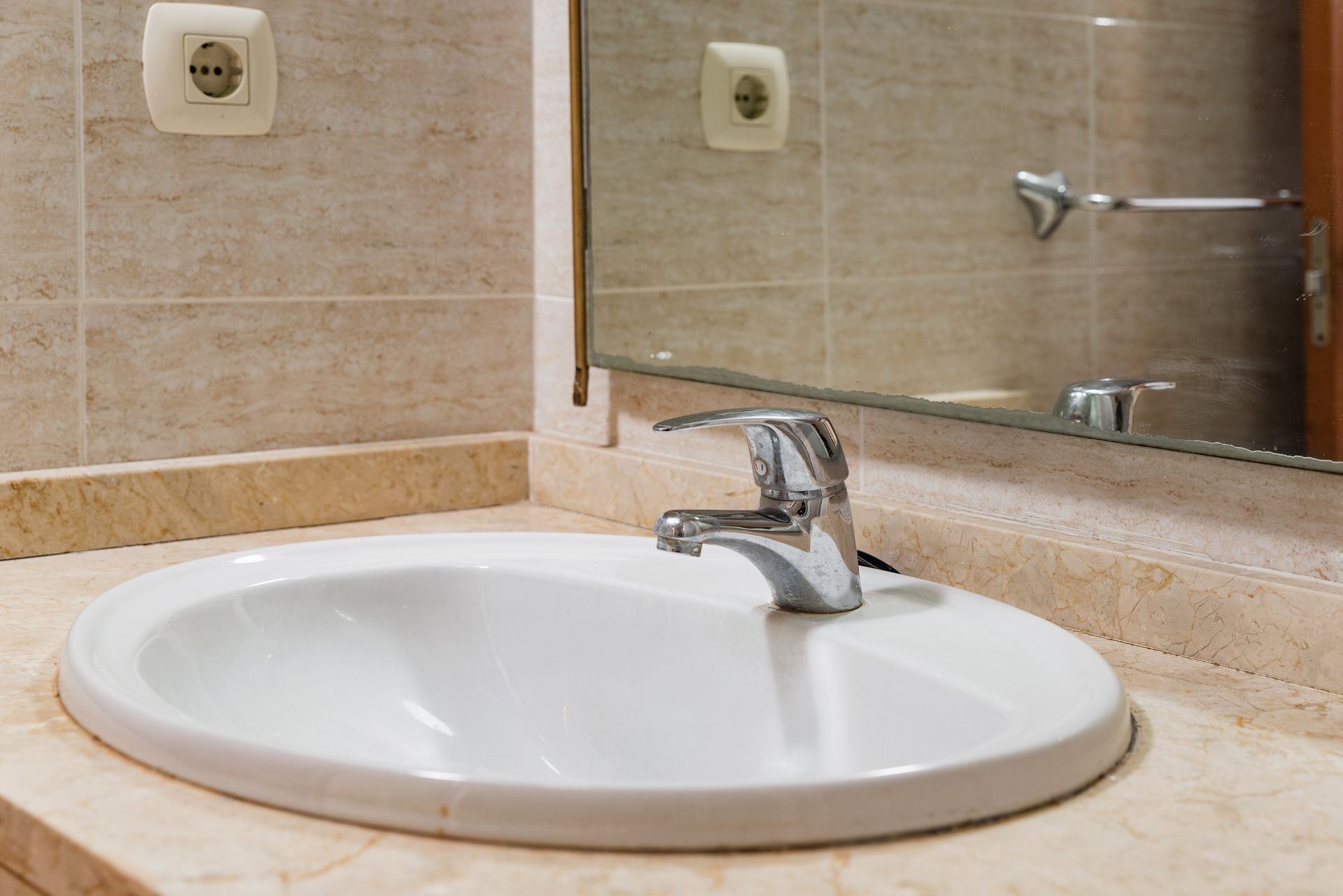 White oval bathroom sink with chrome faucet, set in a marble countertop, with a mirror and wall outlet above.