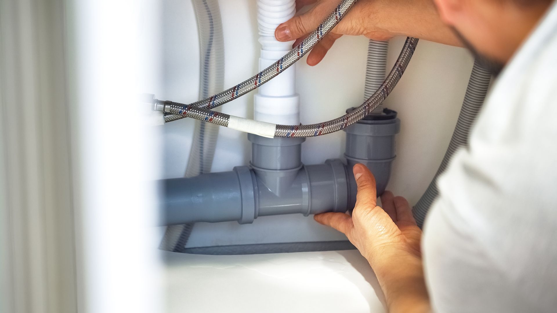 Person installing plumbing under a white sink, working with gray pipes and silver hoses.