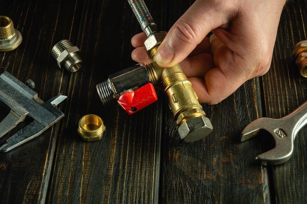 A hand assembling plumbing parts on a dark wooden surface. Tools and brass fittings are scattered around a red-handled valve.