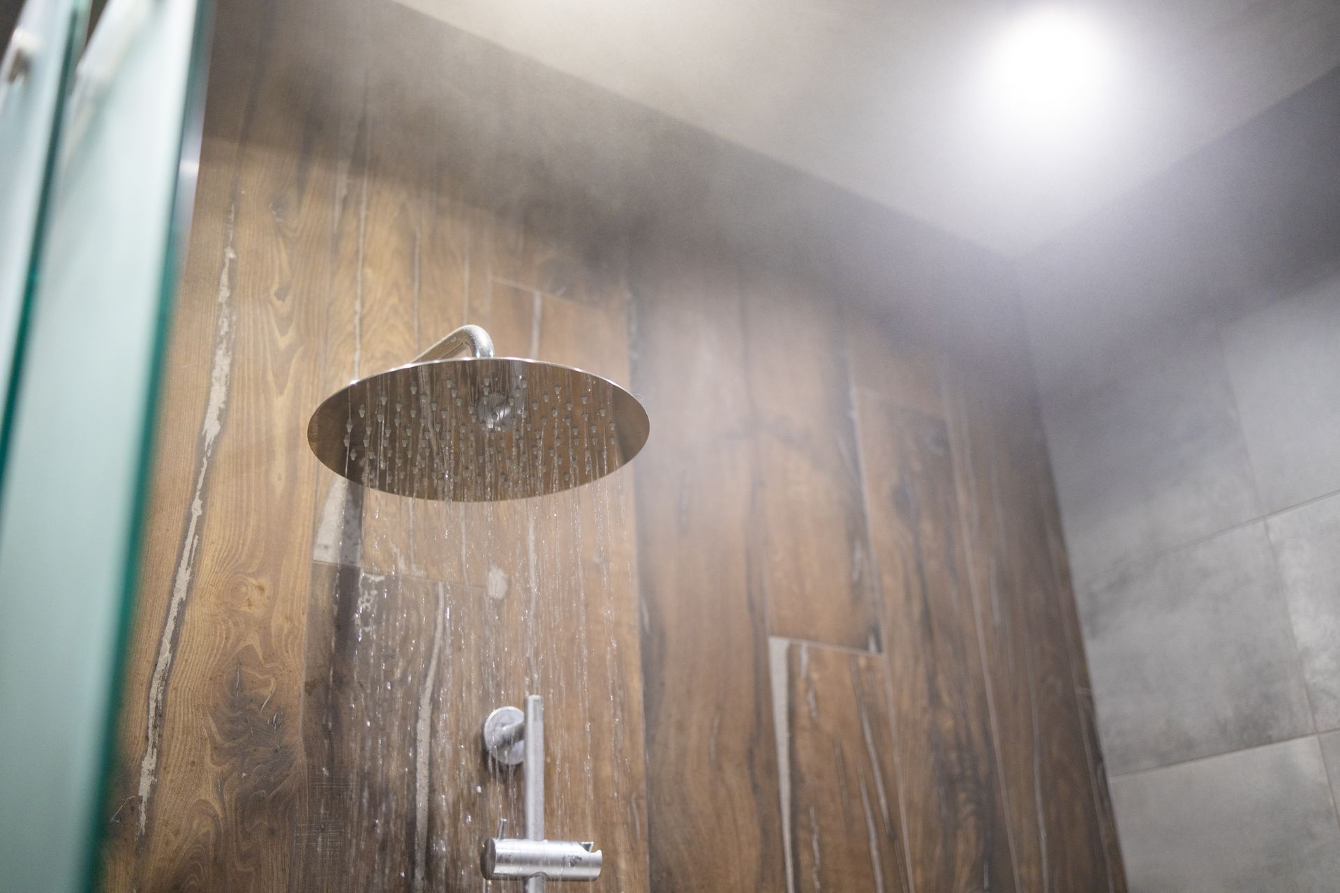 Shower head with water spraying, steam present against wood panel wall.