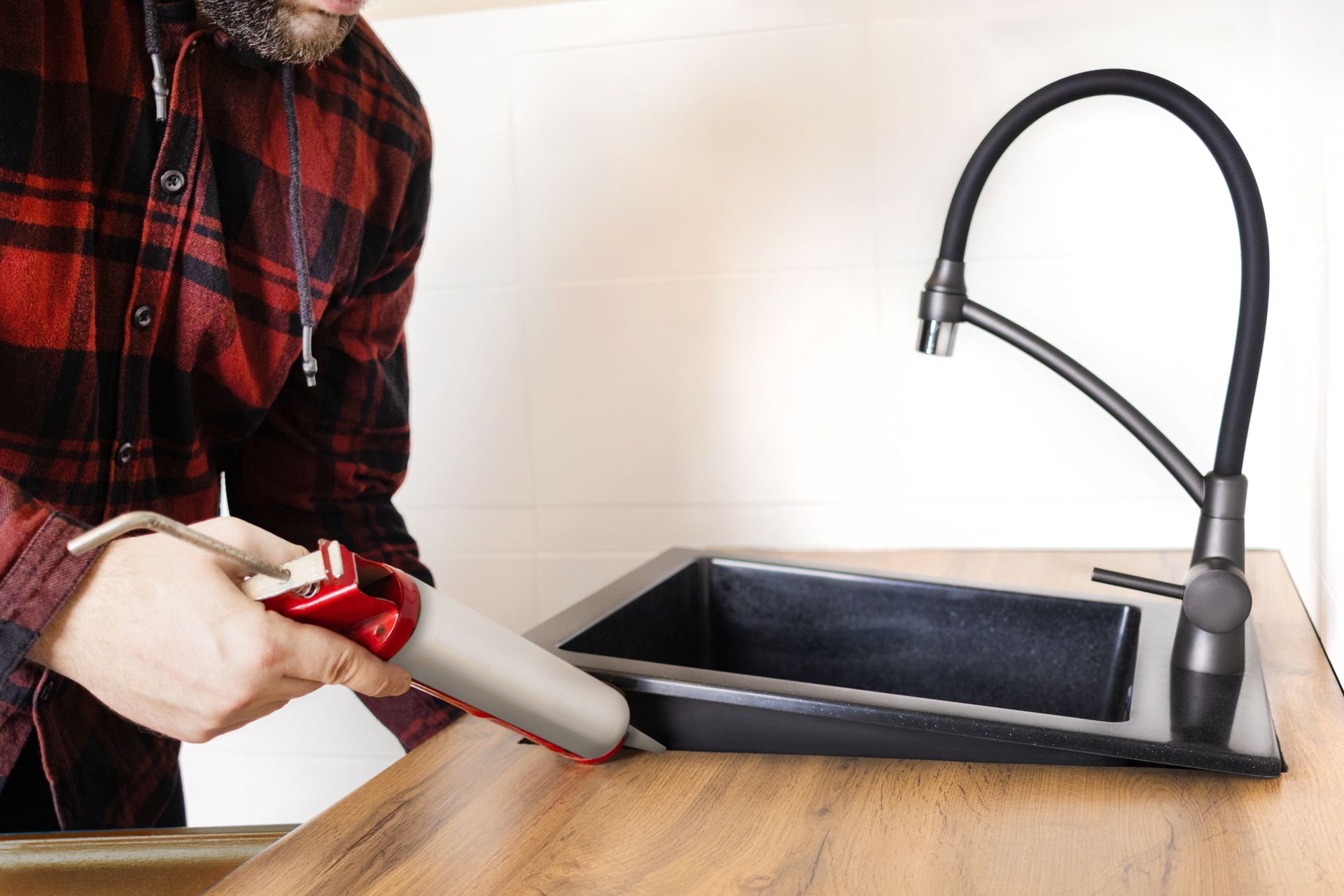 Man caulking around a black sink in a wooden countertop, wearing a red plaid shirt.
