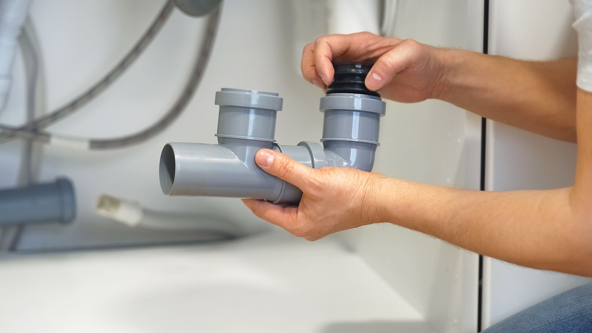 Hands assembling a gray PVC sink drain pipe under a white sink.