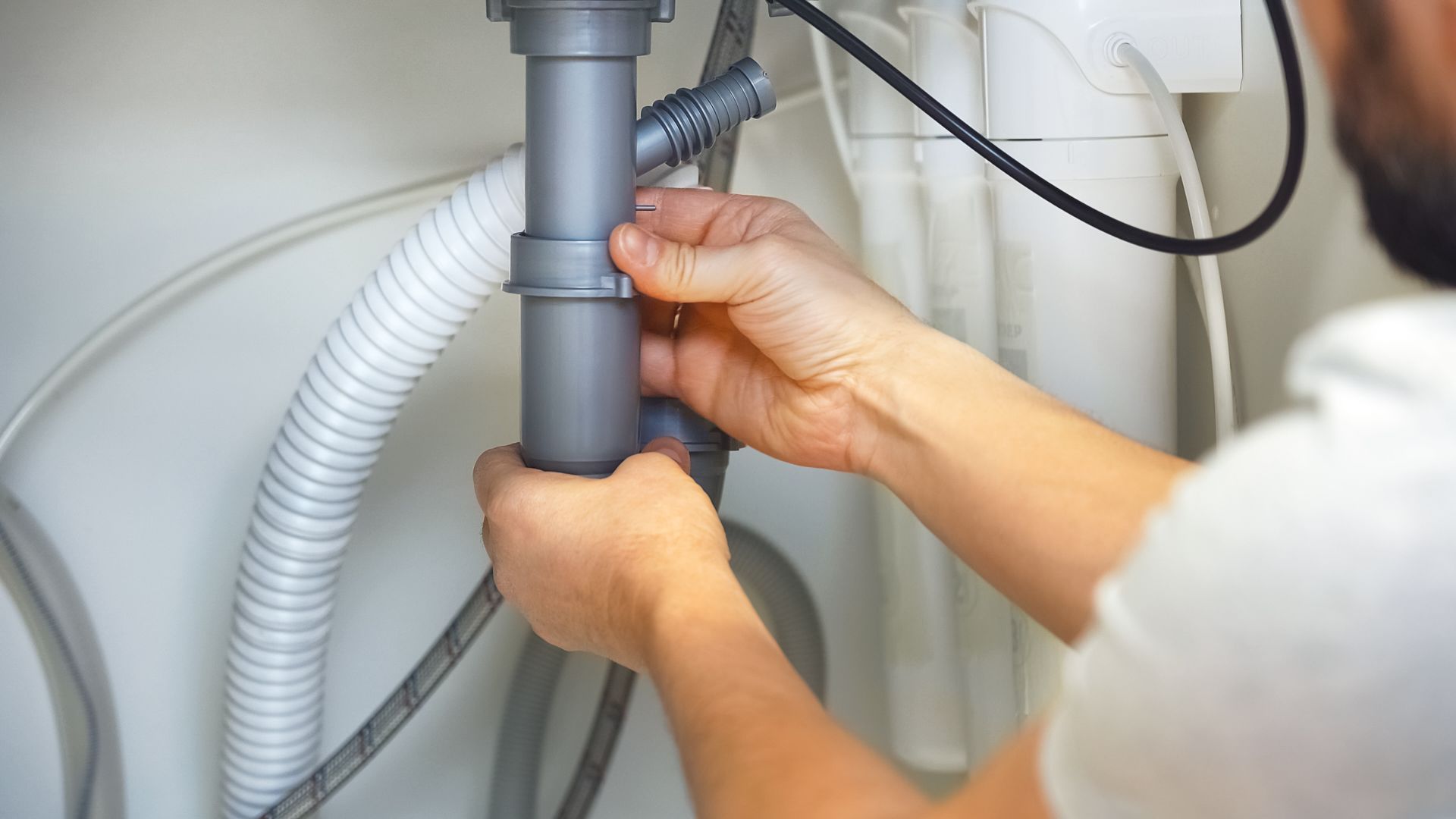 Person assembling plumbing pipes under a sink; hands near a gray pipe.