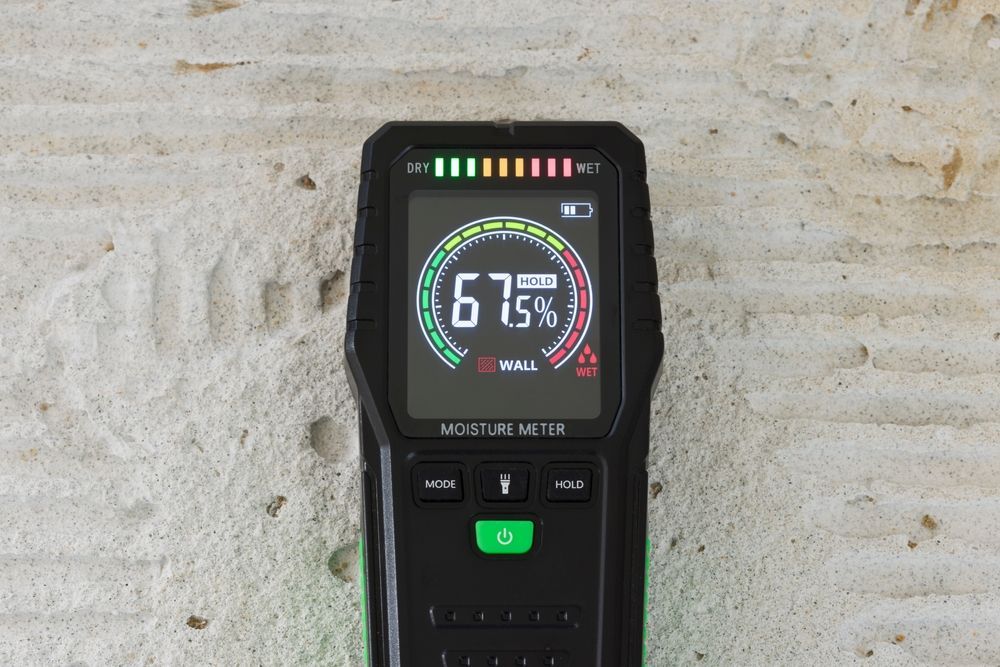 A moisture meter displays 67% on a wall, with a green to red bar graph, indicating high moisture.