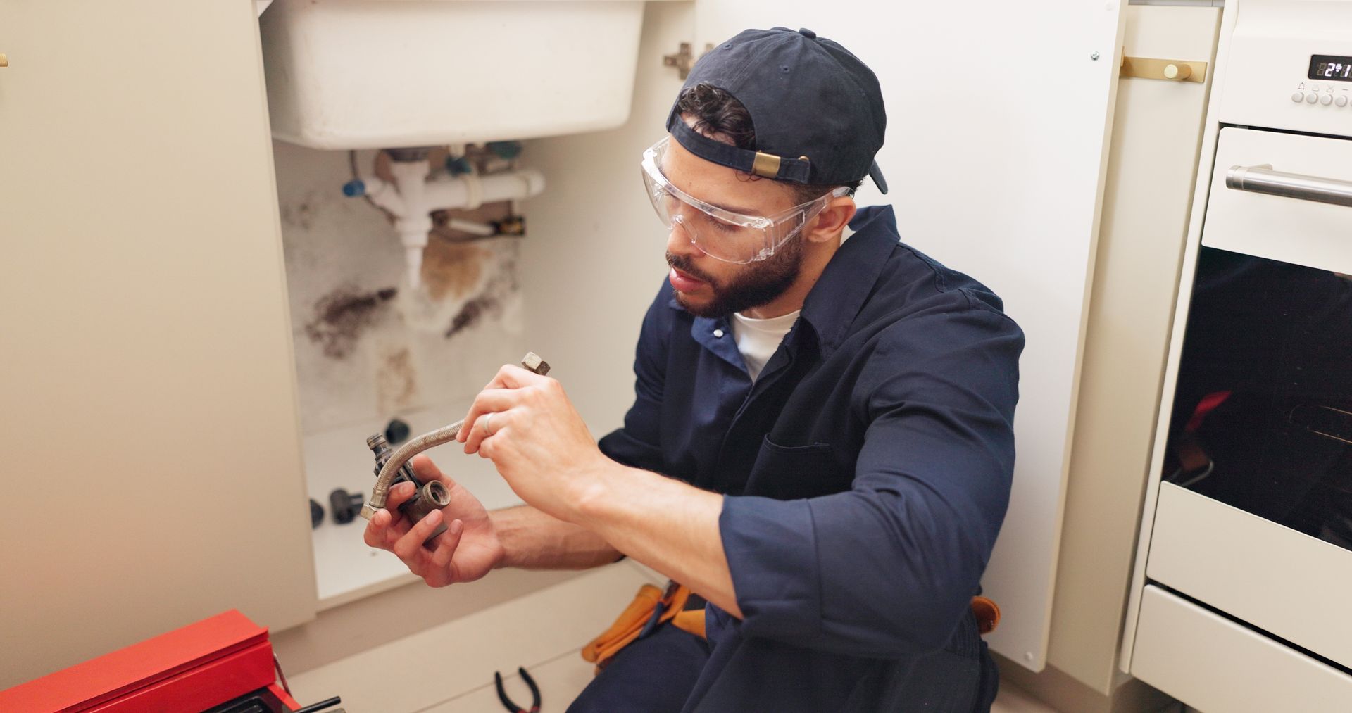 Plumber working under a kitchen sink, wearing safety glasses and a cap, repairing pipes.