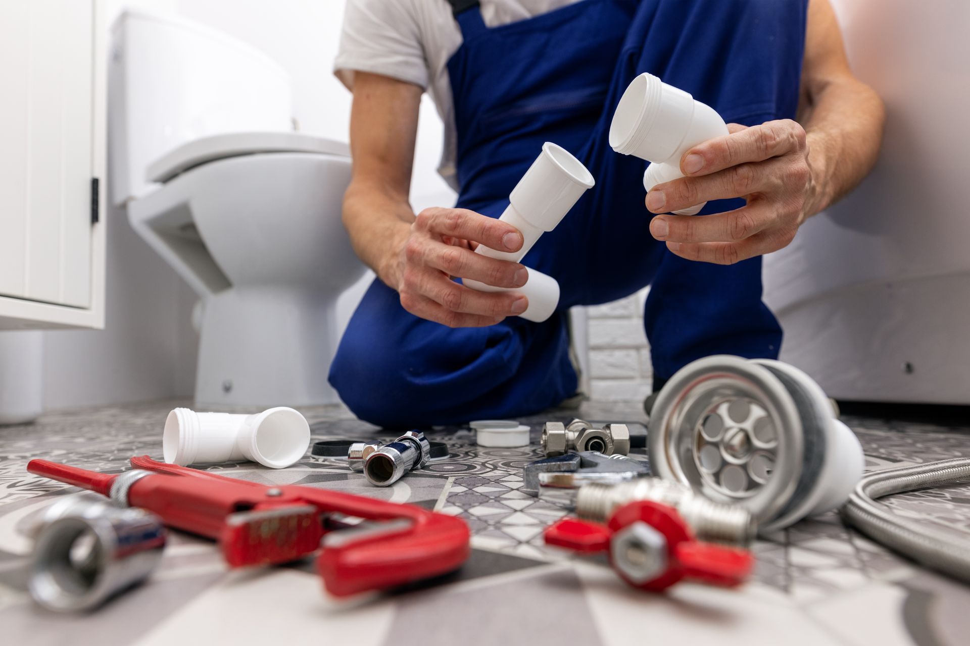 Plumber in blue overalls kneeling, holding white pipes, surrounded by tools near a toilet.