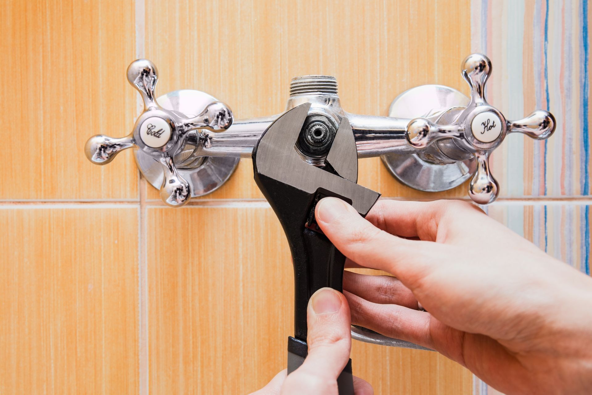 Hands using a wrench to repair a chrome shower faucet on a tiled wall.