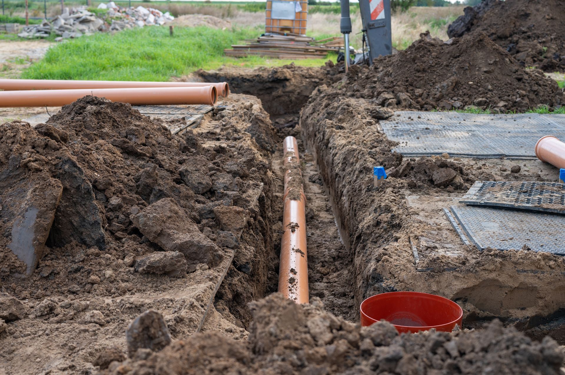 A trench with brown pipes installed, surrounded by dirt and construction materials outdoors.