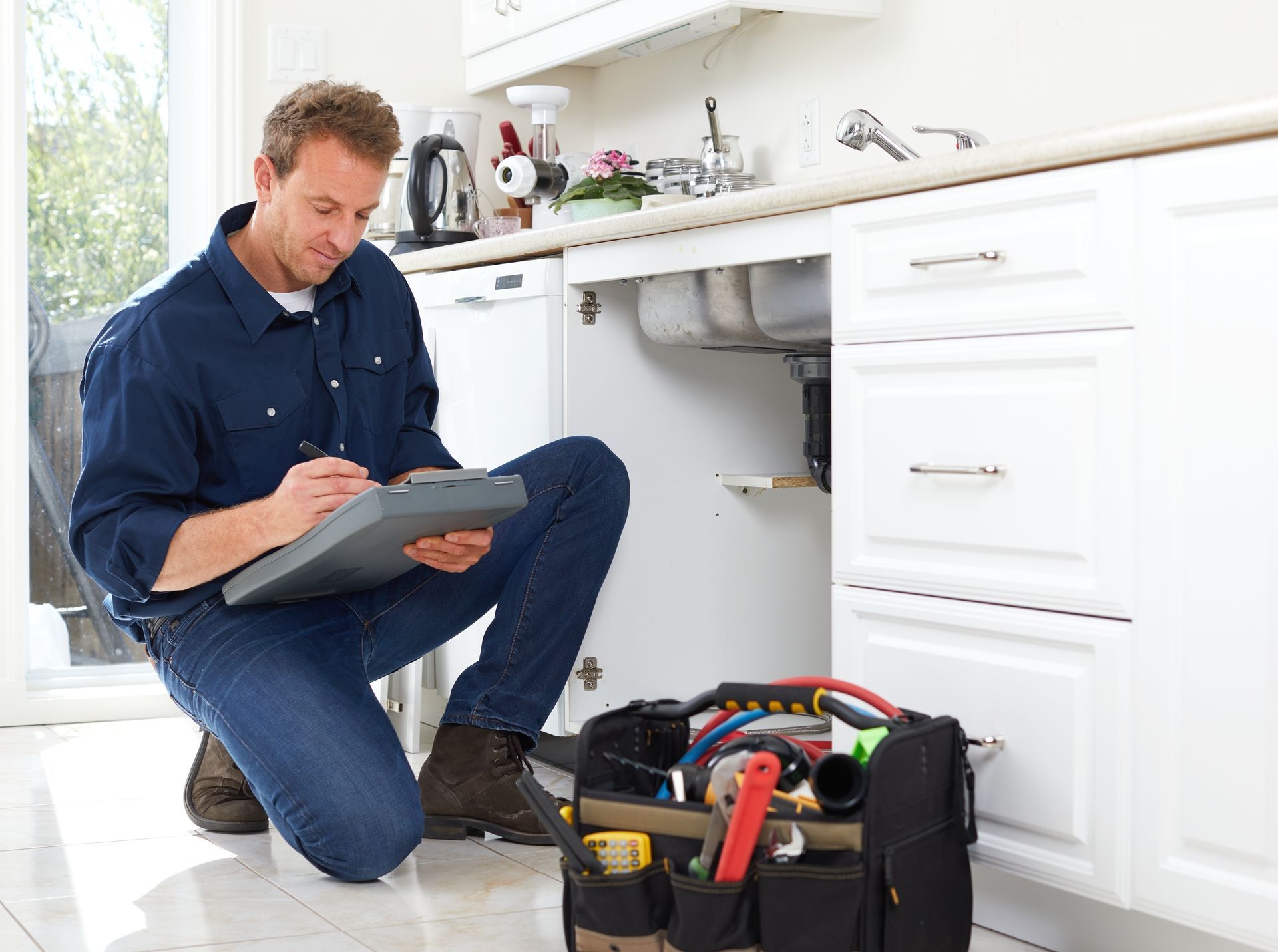 Plumber kneeling at a kitchen sink, writing on a clipboard, toolbox beside him.