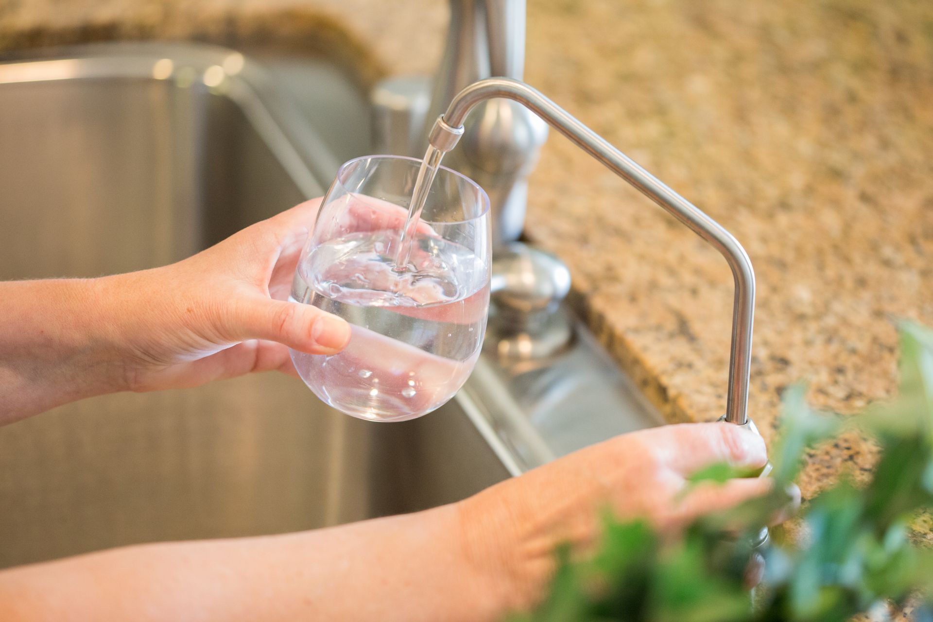 Person filling a glass with water from a kitchen faucet, next to a stainless steel sink.