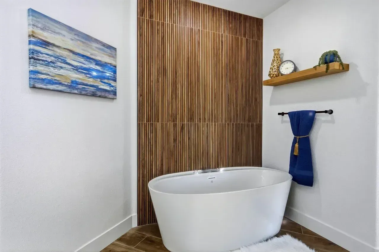 Bathroom with white tub, wood accent wall, blue artwork, and floating shelf.