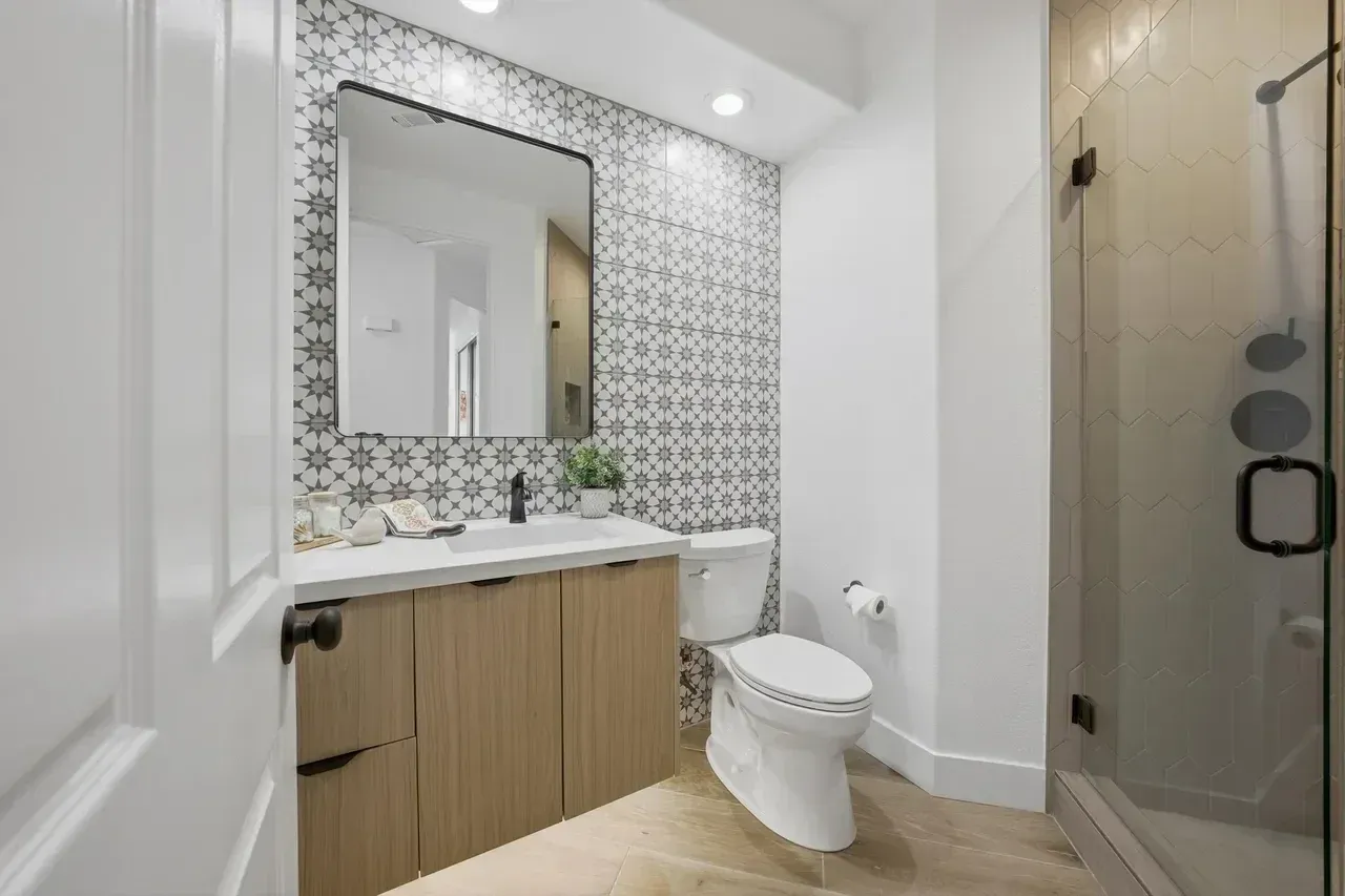 Bathroom with patterned tile accent wall, light wood vanity, toilet, and glass shower.