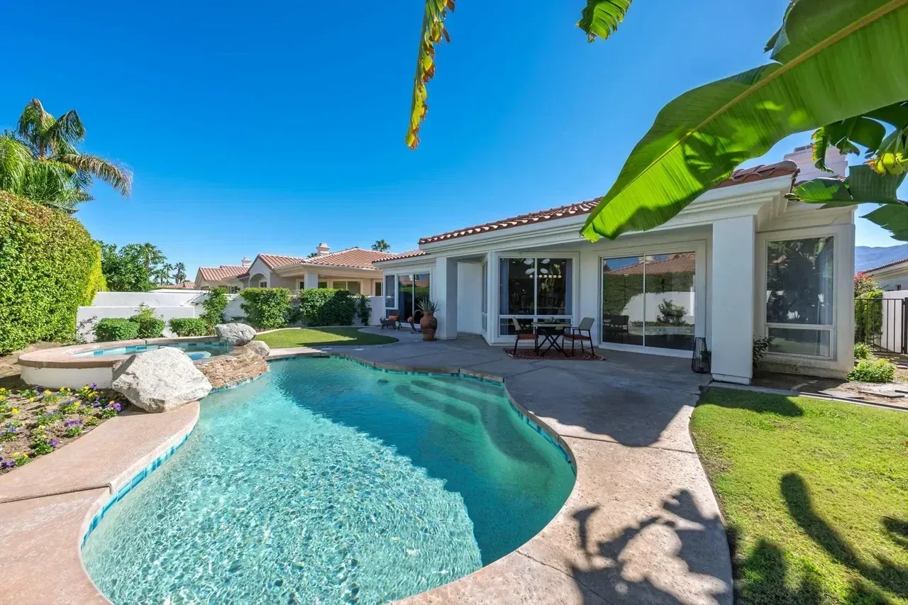 Pool and patio of a home with blue water, green grass, and sunny sky.