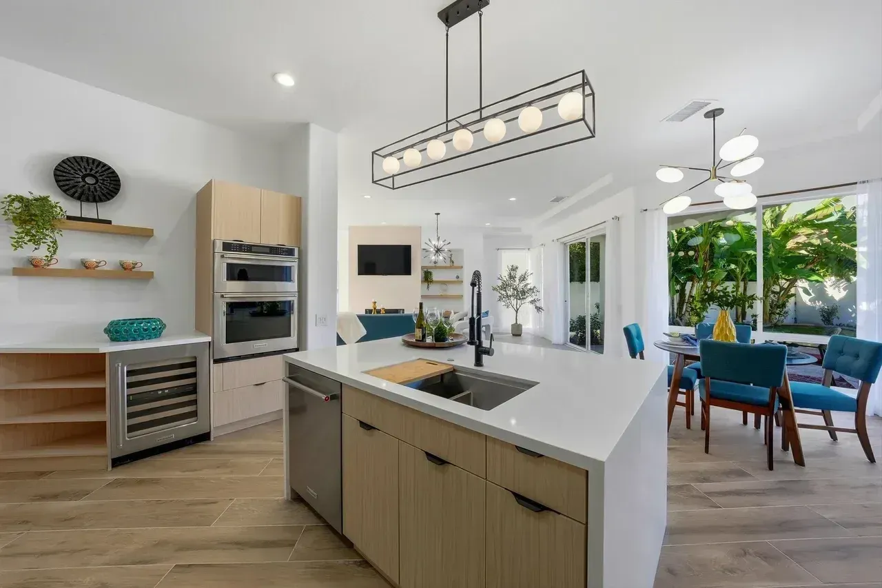 Modern kitchen with light wood cabinets, white countertops, and an island; dining area with blue chairs and lush plants.