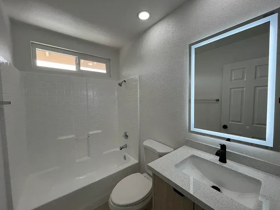 White bathroom with a tub, toilet, vanity, and a lit-up mirror.