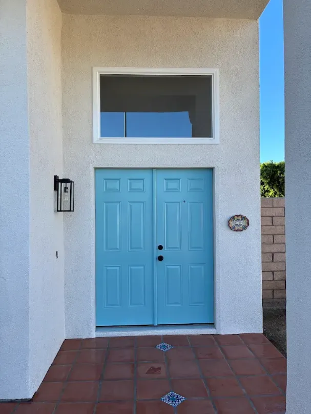 Bright blue double doors with a window above, set in a white stucco entryway with terracotta tile flooring.