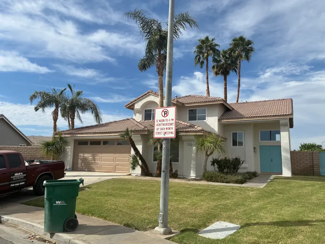 Two-story house with tan garage door, light blue front door, palm trees, blue sky, no parking sign.
