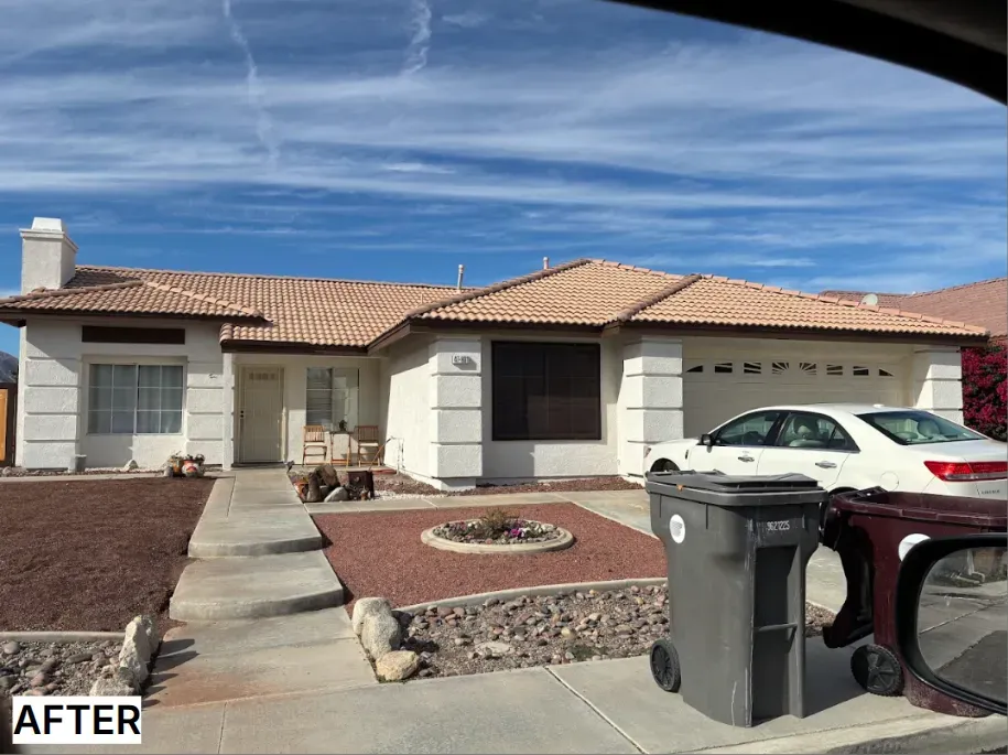 White house with brown roof and a white car in the driveway, with trash bins in front.