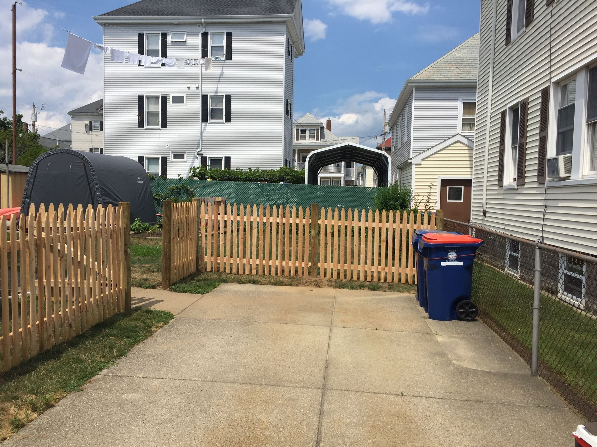 A wooden picket fence surrounds a driveway between two houses.