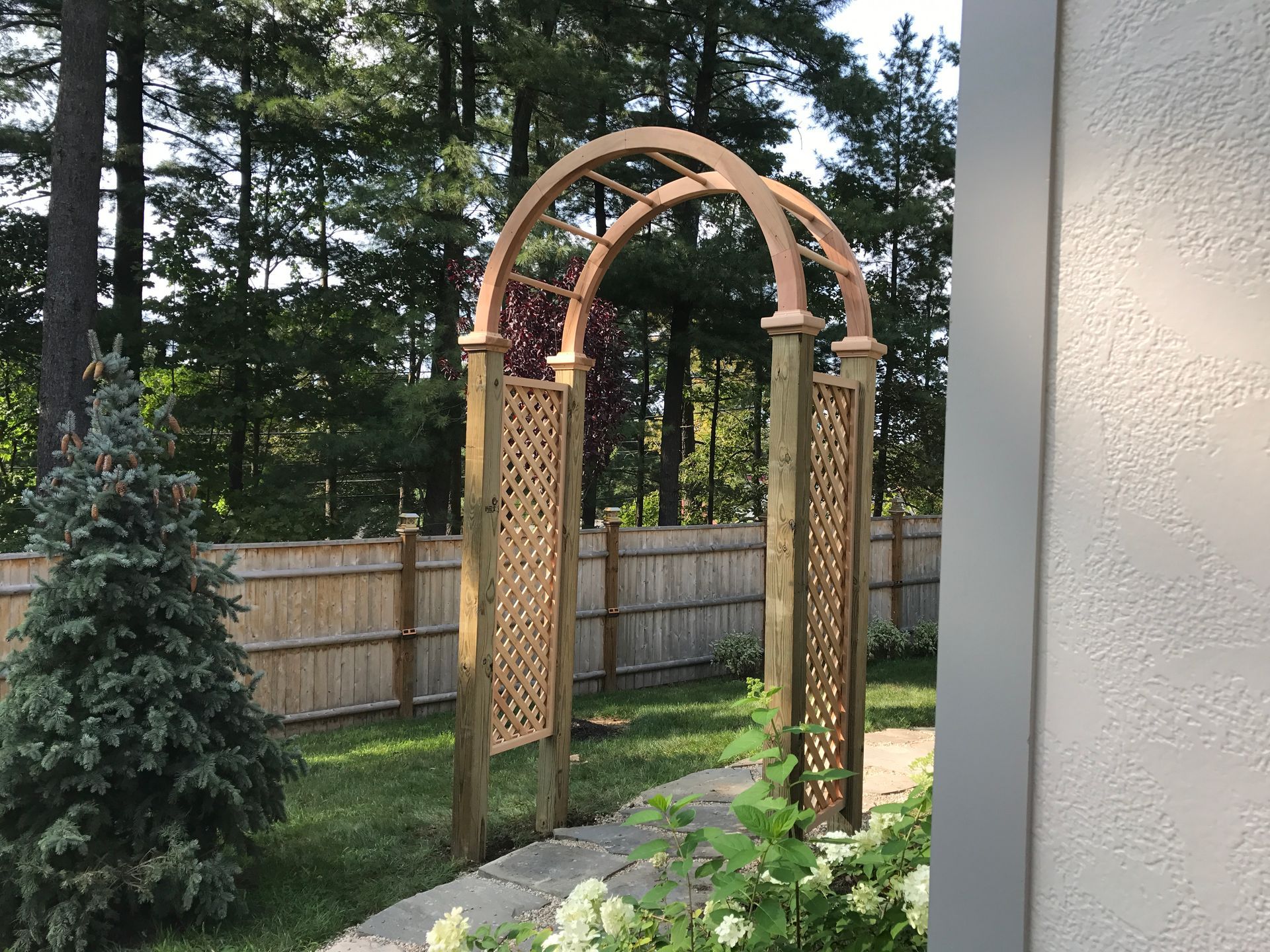 A wooden archway in a backyard with a fence and trees in the background.