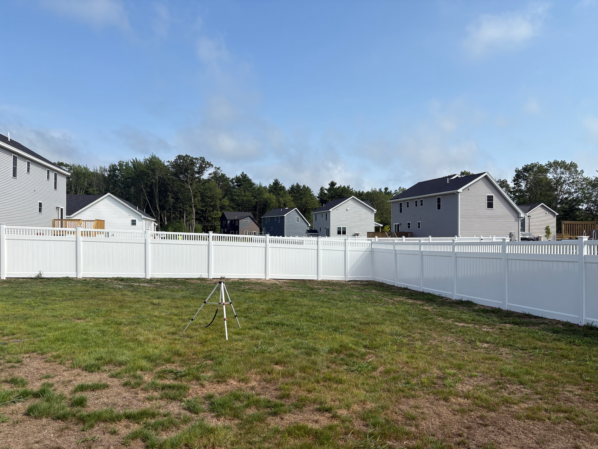A white fence surrounds a lush green yard with houses in the background.
