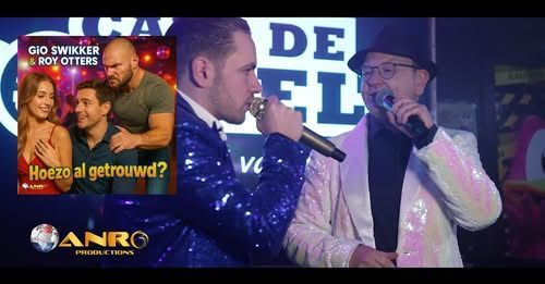 Twee mannen zingen met microfoons op een podium; een videofragment toont een stel en een gespierde man.