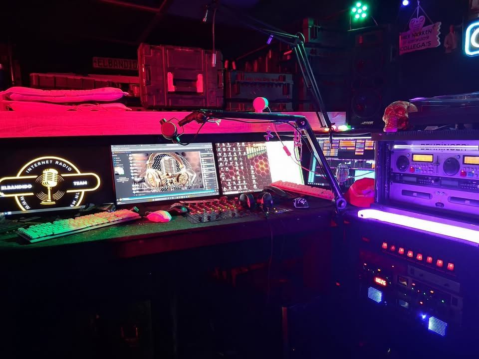 Donkere ruimte met uitzendapparatuur: meerdere monitoren, audioapparatuur en neonverlichting.